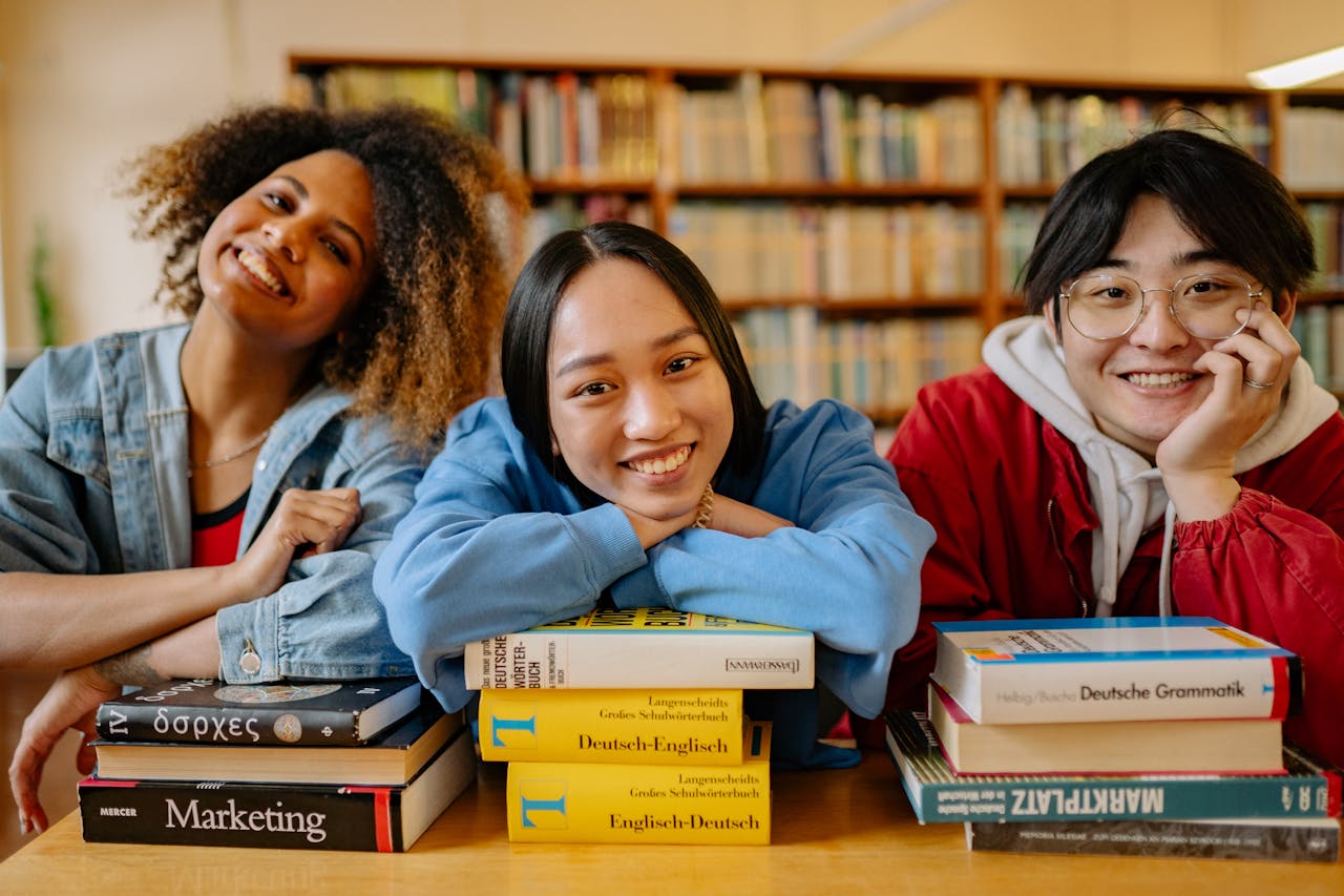 Three smiling students in a library, engaged in studying with stacks of books.